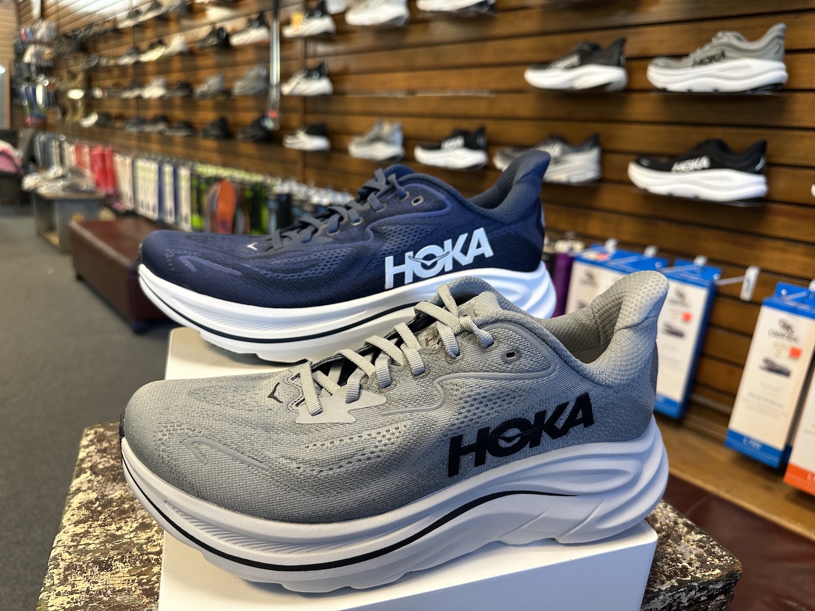 A pair of HOKA running shoes displayed in a store, one in navy blue and the other in gray, with a collection of various shoes visible on the wall in the background.